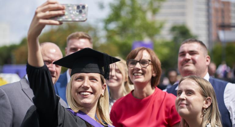 A graduate in a black robe and cap with a purple sash taking a selfie with family members