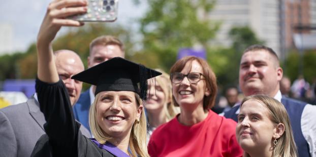 A graduate in a black robe and cap with a purple sash taking a selfie with family members