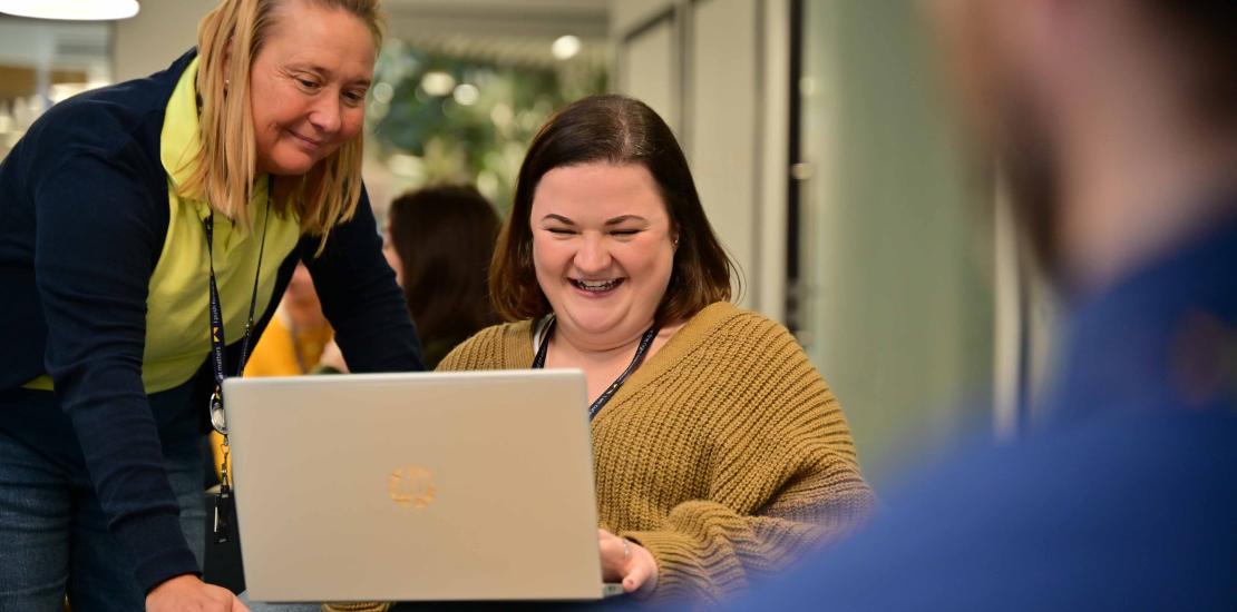 Two colleagues, one seated and one standing, look at a laptop together
